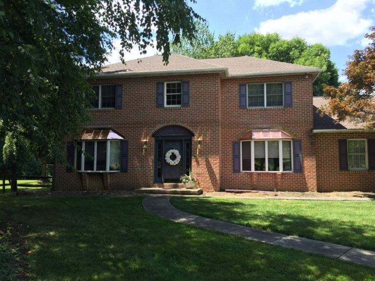 red brick house in East Petersburg PA with blue shutters and a blue door and a concrete path leading to house through grass shrouded by tree